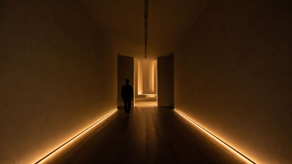 Dim stone-and-wood threshold corridor at evening, lit only by a low cove glow at the floor and a faint warm pool at the far end where the corridor opens into a brighter hall.